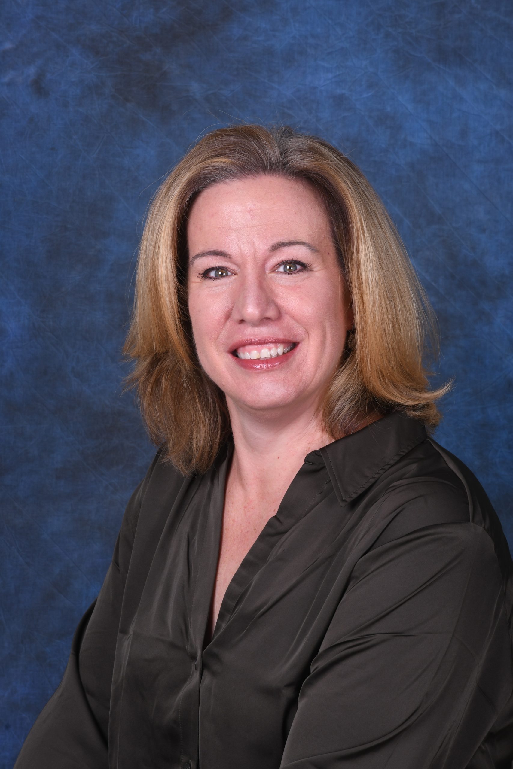 Business headshot of a woman with shoulder-length blonde hair in a dark olive satin blouse, smiling at the camera against a mottled blue painted backdrop.